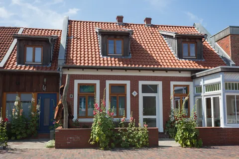 Straßenansicht des Vormannshuus auf Borkum: Klinkerhaus mit roten Dachziegeln, Gauben und Stockrosen vor dem Eingang